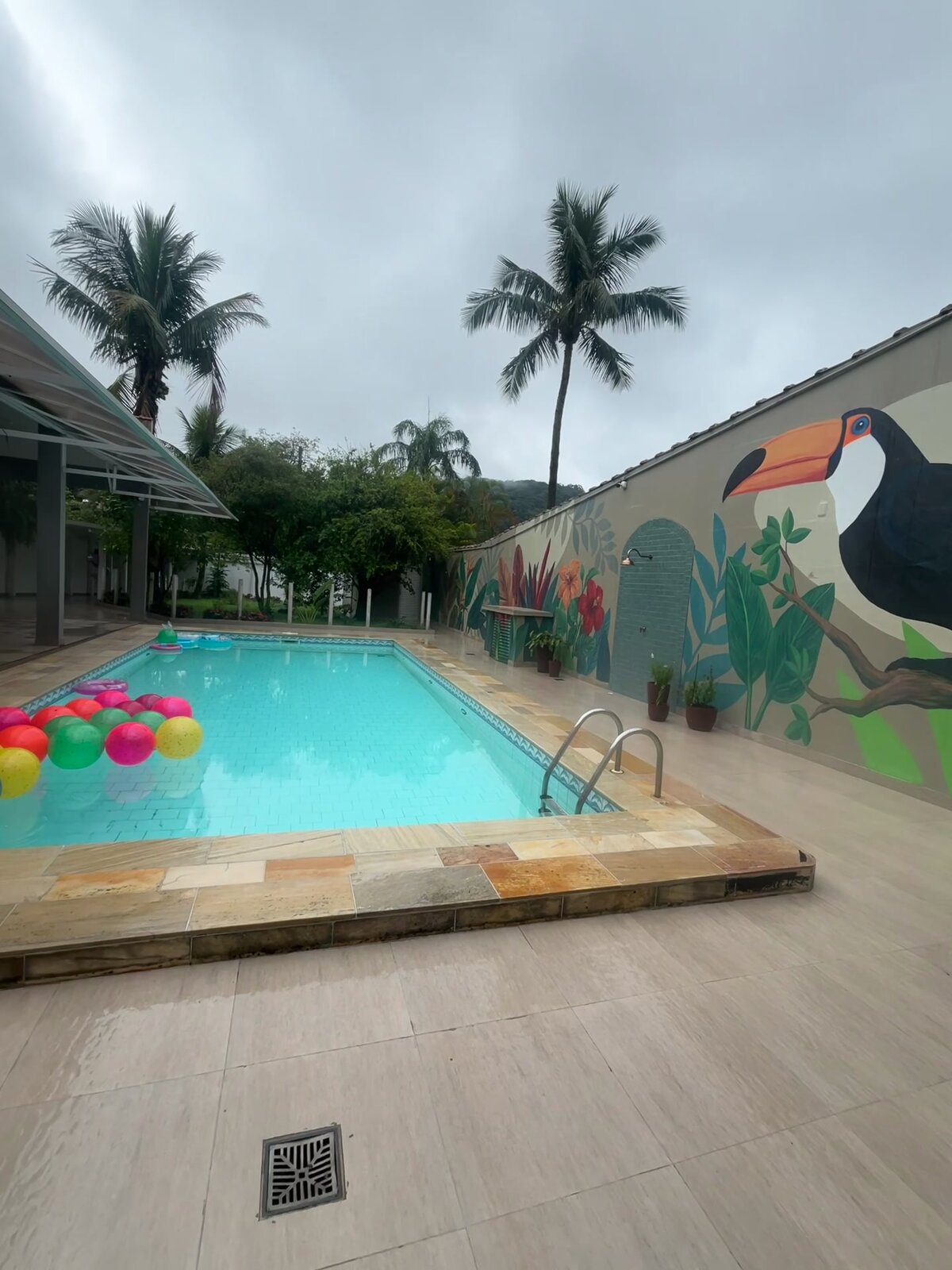 Piscina da Casa 2 com bolas coloridas na água, o mural do tucano e as palmeiras atrás.